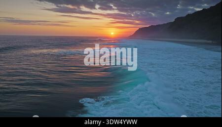 Coucher de soleil doré sur la côte de l'océan Indien, ciel de nuages colorés lumineux se reflète sur la vaste surface de l'eau, vagues puissantes brisent la côte tropicale de l'île de Bali avec des falaises vertes luxuriantes en arrière-plan. Prise de vue aérienne Banque D'Images