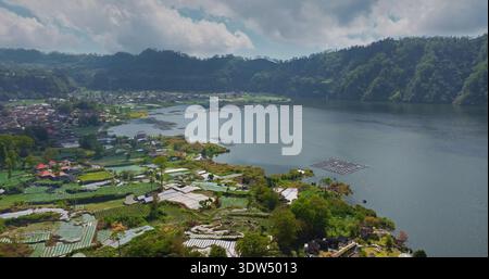 Indonésie, Bali, Abang : vue aérienne panoramique sur le lac Batur avec ses fermes piscicoles flottantes, les villages environnants et de vastes terrasses agricoles sur les pentes volcaniques fertiles. Vol aérien par drone Banque D'Images