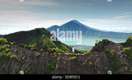 Homme célébrant le succès sur la crête de montagne avec les bras tendus, profitant d'une vue panoramique sur la caldeira, le lac Batur et le mont Agung au loin, représentant la réalisation et la liberté. Vol aérien Banque D'Images