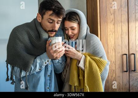 Joyeux jeune couple couvert de couverture embrassant tout en tenant des tasses de thé à la maison Banque D'Images