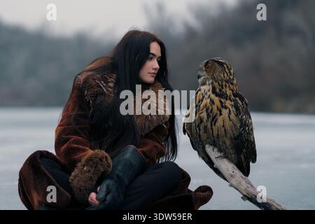 Jeune femme en manteau de fourrure assise sur le lac gelé avec hibou aigle eurasien perché sur la branche. Archer femelle et son compagnon hibou en plein air dans un sauvage hivernal Banque D'Images