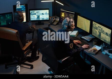 Femme caucasienne d'âge moyen et homme noir surveillant plusieurs écrans de surveillance dans la salle de contrôle de sécurité, tous deux concentrés sur l'observation des flux vidéo en direct et l'analyse des informations sur les moniteurs Banque D'Images
