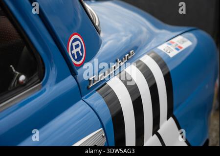 Gros plan de l'emblème chromé Dauphine et d'un logo Renault circulaire sur la carrosserie latérale bleue d'une voiture classique française avec une rue de course noire et blanche Banque D'Images