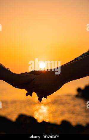 Gros plan de deux personnes se tenant la main au lever du soleil au bord de la mer Banque D'Images