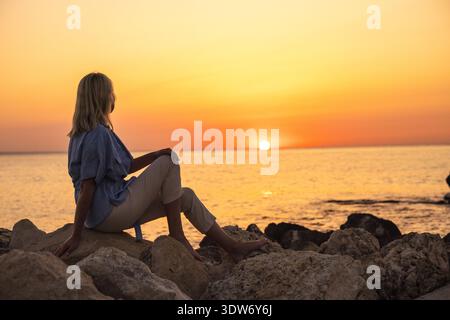 Femme d'âge moyen assise sur les rochers côtiers au coucher du soleil Banque D'Images