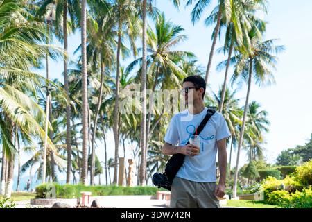 Jeune homme en T-shirt blanc et lunettes de soleil tenant du café glacé marchant à travers Tropical Palm Tree Park en plein soleil avec profondeur de champ cinématographique Banque D'Images