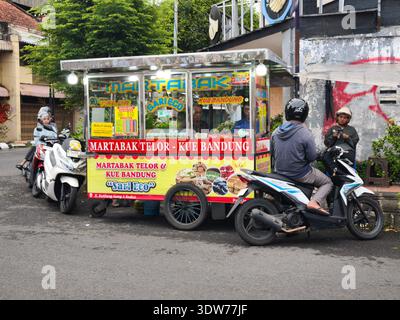 Une charrette de nourriture de rue indonésienne dynamique vendant Martabak et Kue Bandung est garée au bord de la route, entourée de motocyclistes qui attendent leur commande. Banque D'Images