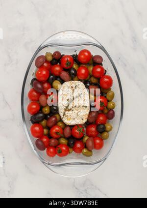 Fromage blanc (camembert, brie) à cuire avec tomates cerises, olives noires et vertes, kalamata, thym et huile d'olive dans un bol en verre sur un dos en marbre Banque D'Images