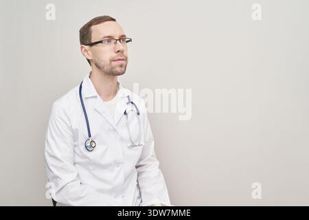 Médecin masculin caucasien en manteau blanc avec stéthoscope regardant avec confiance sur le côté dans le cadre médical professionnel. Banque D'Images