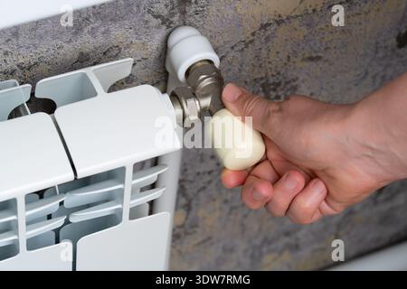Une main humaine tourne la poignée de la vanne sur un radiateur blanc, régulant la température dans le système dans la pièce. Banque D'Images