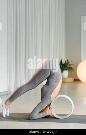 Une femme effectue une pose de yoga en utilisant une roue pour le soutien dans un studio rempli de lumière. L'arrière-plan présente un décor simple avec des plantes et un éclairage doux. Banque D'Images