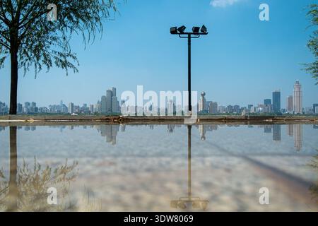 Plan à angle bas d'une flaque de pluie sur le sol reflétant le ciel, avec un haut lampadaire et la ligne d'horizon lointaine de la ville visible à l'horizon. Banque D'Images
