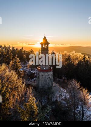 La tour de guet dans les montagnes de Jizera se dresse haute lorsque le soleil se couche. Des arbres entourent la tour, et le paysage hivernal brille de lumière dorée alors que le jour se transforme en nuit. Banque D'Images