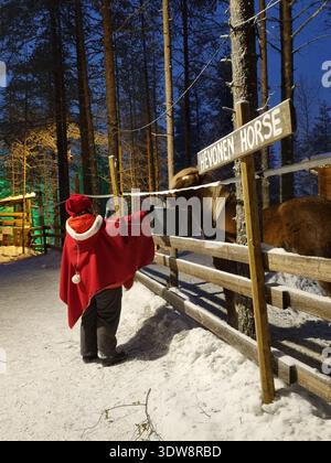 Femme dans Red Cape Feeding Horse dans le village du Père Noël, Rovaniemi Finlande - soirée d'hiver magique en Laponie Banque D'Images
