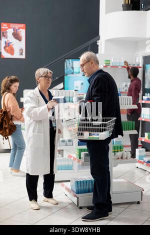 Femme pharmacienne se tient à côté du client masculin âgé, expliquant les options de soins de santé près des étagères du magasin. L’interaction met en valeur la confiance, l’expertise et le service personnalisé dans une pharmacie propre et moderne. Banque D'Images