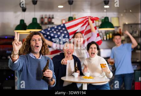 Groupe d'amis supporters acclamant pour l'équipe préférée avec drapeau des États-Unis tout en regardant match à la télévision dans le pub Banque D'Images