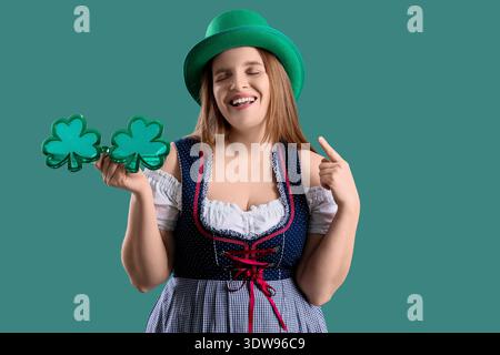 Serveuse irlandaise rousse avec des lunettes en forme de trèfle pointant vers le chapeau de leprechaun sur fond vert. Célébration de la fête de Patrick Banque D'Images