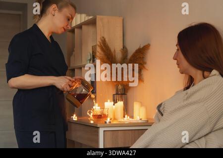 Femme thérapeute versant du thé chaud d'une théière en verre dans une tasse, offrant une cérémonie du thé apaisante dans un environnement de spa avec des bougies Banque D'Images