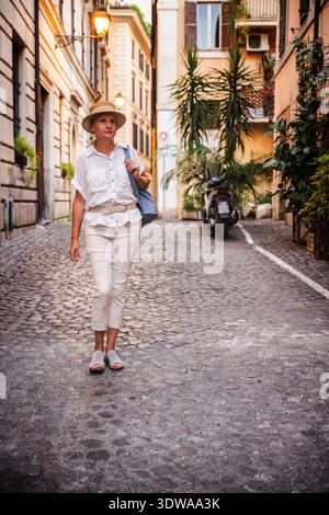 Femme d'âge moyen portant un chapeau de paille et des vêtements légers marchant avec un sac à dos le long d'une rue pavée de Rome entourée de bâtiments historiques Banque D'Images