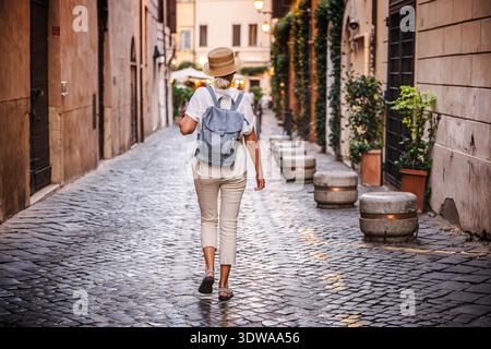 Femme d'âge moyen avec chapeau de paille et sac à dos marchant le long d'une rue pavée de Rome avec des lumières du soir et de charmants bâtiments historiques Banque D'Images