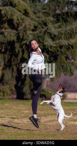 Une jeune femme joyeuse saute et danse avec son chien ludique dans un parc ensoleillé, profitant de l'exercice en plein air et se liant avec son animal énergique. Banque D'Images