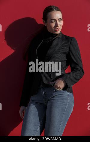 Une femme se tient confiante contre un mur rouge vif, vêtue d'un blazer noir et d'un Jean bleu. Banque D'Images