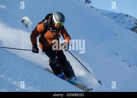 Un skieur vêtu d'une veste orange, portant un casque et des lunettes de protection, sculpte à travers la poudre fraîche sur une pente de montagne enneigée. La scène transmet la vitesse, la mise au point et l'aventure hivernale en plein air. || autorisation du modèle Banque D'Images