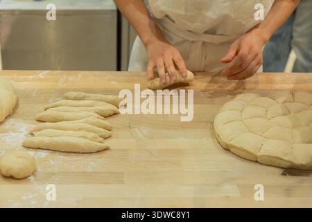 Baker façonne habilement la pâte sur une surface en bois, entourée de différentes formes de pain et de farine, mettant en valeur l'art culinaire. Banque D'Images