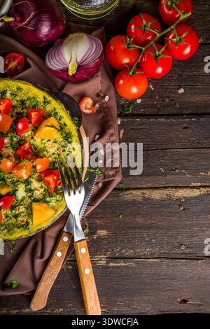 Frittata maison de style rustique. Frittata aux épinards, au fromage et à la tomate dans une poêle. Arrière-plan en bois avec espace de copie de légumes Banque D'Images