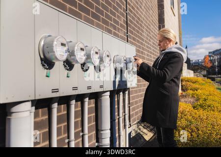 Femme vérifiant les compteurs électriques extérieurs sur le mur du bâtiment dans le quartier résidentiel Banque D'Images