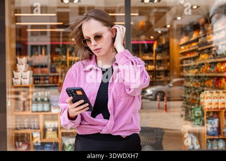 Une jeune femme en chemise rose se tient calmement devant un magasin, concentrée sur son smartphone. La toile de fond urbaine présente une scène urbaine dynamique remplie de b Banque D'Images