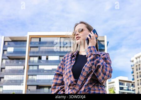 Une femme portant des lunettes de soleil parle sur son téléphone à l'extérieur, dans le contexte de l'architecture urbaine moderne. Le ciel est clair, ajoutant à l'éclatant, vivant Banque D'Images