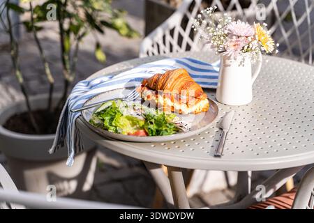 Un délicieux croissant de saumon accompagné d'une salade fraîche est servi sur une table de café ensoleillée. La scène capture un cadre parfait pour une appétizine saine Banque D'Images