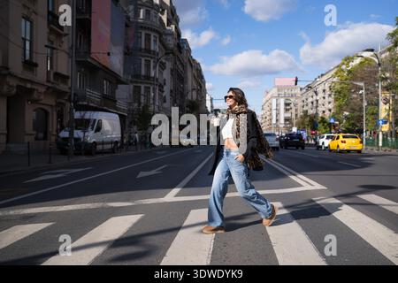 Une femme élégante portant des lunettes de soleil et une écharpe à imprimé léopard se promène décontractée dans une rue en tenant une tasse de café, sur un dos urbain Banque D'Images