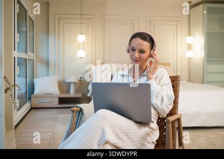 Une jeune femme se détend à la maison, portant des écouteurs et utilisant un ordinateur portable. Elle semble confortable et concentrée, assise dans une chaise confortable avec un mode lumineux Banque D'Images