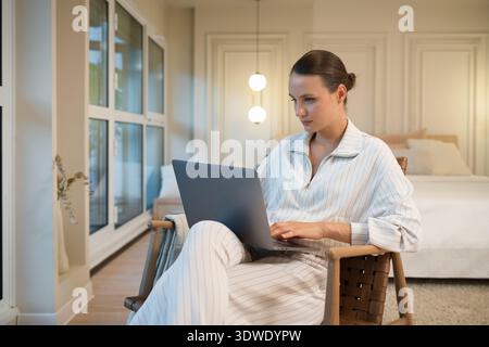Une jeune femme est confortablement assise à la maison, concentrée sur son ordinateur portable. L'éclairage ambiant et le décor moderne créent une atmosphère chaleureuse dans laquelle elle s'engage Banque D'Images