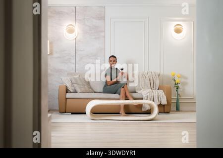 Une jeune femme est assise confortablement sur un canapé moderne, tenant un verre de vin dans un salon élégamment décoré, créant une maison relaxante et sereine à Banque D'Images