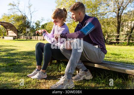 Un couple prend une pause de jogging dans un parc pittoresque, en vérifiant leurs progrès de forme physique sur smartwatches. La lumière du soleil filtre à travers les arbres, soulignant Banque D'Images