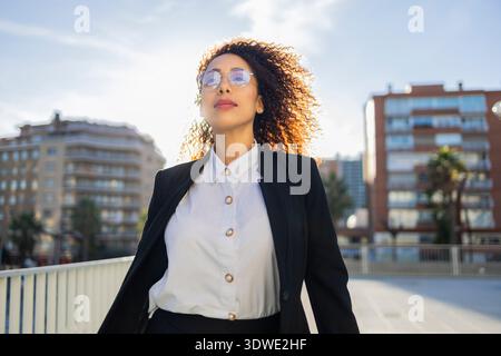 Femme d'affaires avec les cheveux bouclés marchant en toute confiance dans un cadre urbain. Elle est habillée d'un costume professionnel et porte des lunettes, exsudant professionnel Banque D'Images
