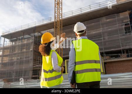 Deux ingénieurs portant des casques de sécurité et des gilets réfléchissants discutent des plans sur un chantier de construction. La structure derrière eux est en cours de développement, présentant des échafoldi Banque D'Images