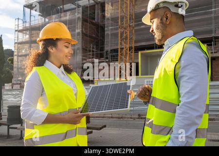 Deux ingénieurs portant des gilets de sécurité et des casques de sécurité discutent d'un panneau solaire sur un site. Ils explorent l'intégration de l'énergie renouvelable dans un projet de construction moderne Banque D'Images