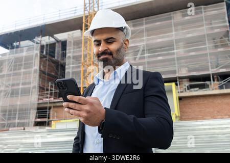 Un ingénieur masculin portant un casque de sécurité et un costume utilise un smartphone sur un chantier de construction, mettant en valeur la technologie moderne dans la gestion de projet au milieu d'un échafo Banque D'Images