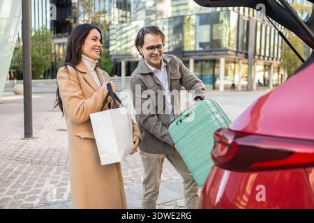 Un couple joyeux charge des bagages dans le coffre de la voiture, se préparant pour un voyage. Ils sont debout à l'extérieur dans un environnement urbain, profitant du moment Banque D'Images