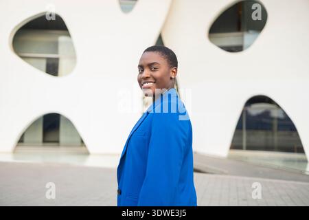 Femme d'affaires africaine confiante dans un costume bleu sourit tout en se tenant à l'extérieur d'un bâtiment moderne. Elle incarne le leadership et le professionnalisme dans une urba Banque D'Images