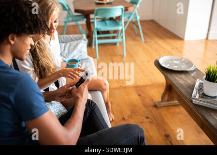 Un couple multiethnique se détend sur un canapé confortable à la maison, interagissant avec un smartphone et buvant du café, illustrant le rôle de la technologie Banque D'Images