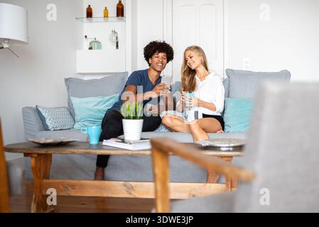 Un couple multiethnique profite d'un après-midi détendu à la maison. Ils sont assis ensemble sur un confortable canapé gris, sirotant un café et regardant un smartph Banque D'Images
