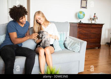 Un couple multiethnique profite d'un moment cosy à la maison avec leur lapin de compagnie. Assis sur un canapé confortable dans un salon bien décoré, ils partagent un Banque D'Images