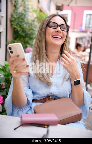 Une femme mature dans un café profitant d'une journée ensoleillée, souriante et tenant un smartphone. Elle porte des lunettes et une smartwatch, créant un a joyeux et détendu Banque D'Images