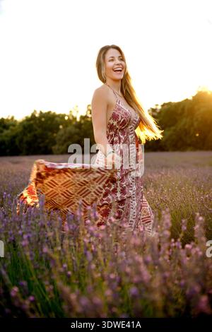 Une femme joyeuse dans une robe à motifs marche à travers un champ de lavande serein au coucher du soleil, avec ses cheveux coulant gracieusement, capturant l'essence de l'été Banque D'Images
