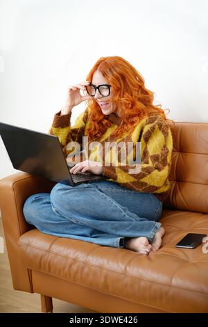 Une femme joyeuse aux cheveux roux et aux lunettes rit en travaillant sur son ordinateur portable. Elle est assise sur un canapé en cuir marron, profitant d'un Mo détendu et heureux Banque D'Images